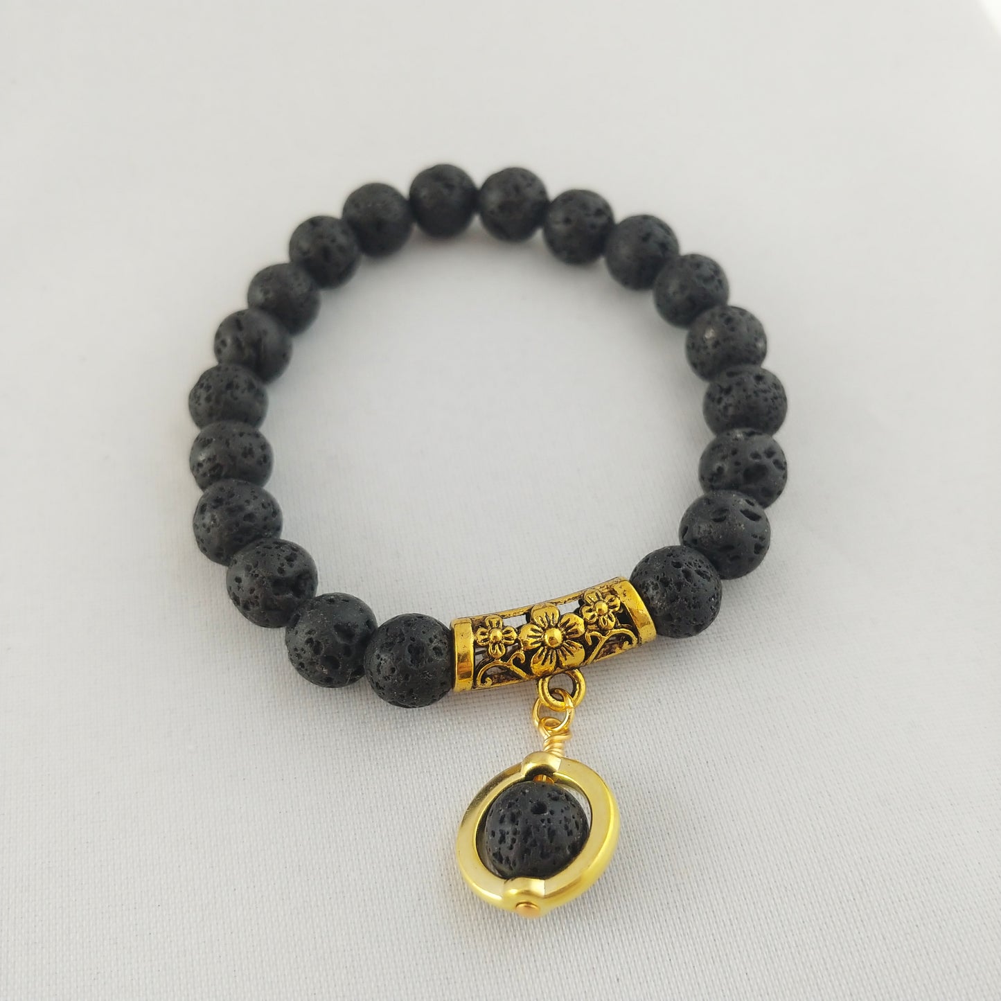 Lava bracelet sitting on a white background. Bracelet has lav astones and a golden flower centerpiece, from that a golden circle is dangling which has a lava stone in the center.
