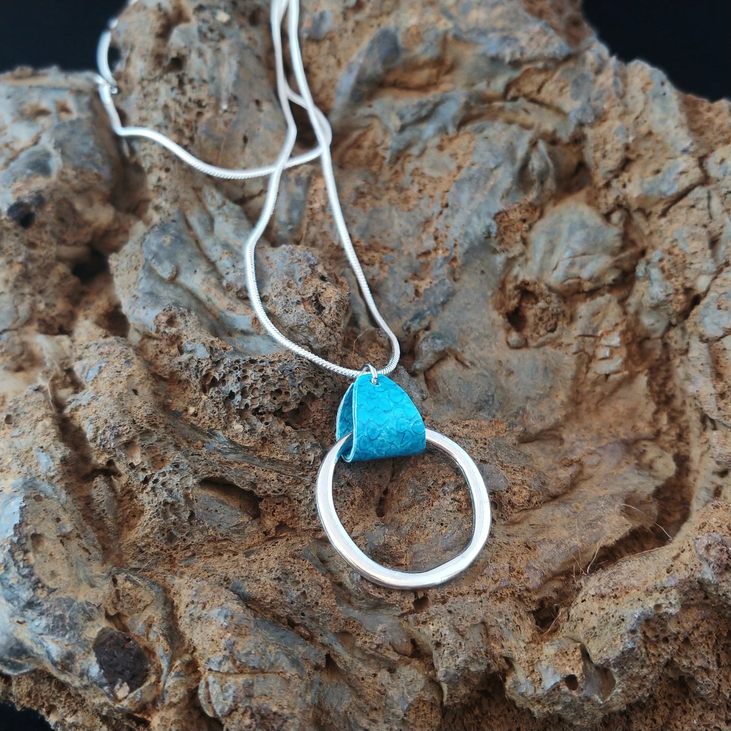 Silver colored necklace with short silver chain and a circle silver center piece. The chain and circle are connected with bright blue Icelandic salmon leather piece.