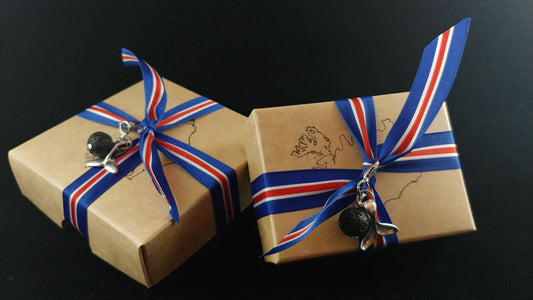 Two gift boxes sitting on a dark black display cloth. The boxes have the outlines of iceland stamped on them and each box is wrapped with an icelandic flag ribbon. On each ribbon there is a small charm with a whale tail and a lava bead.