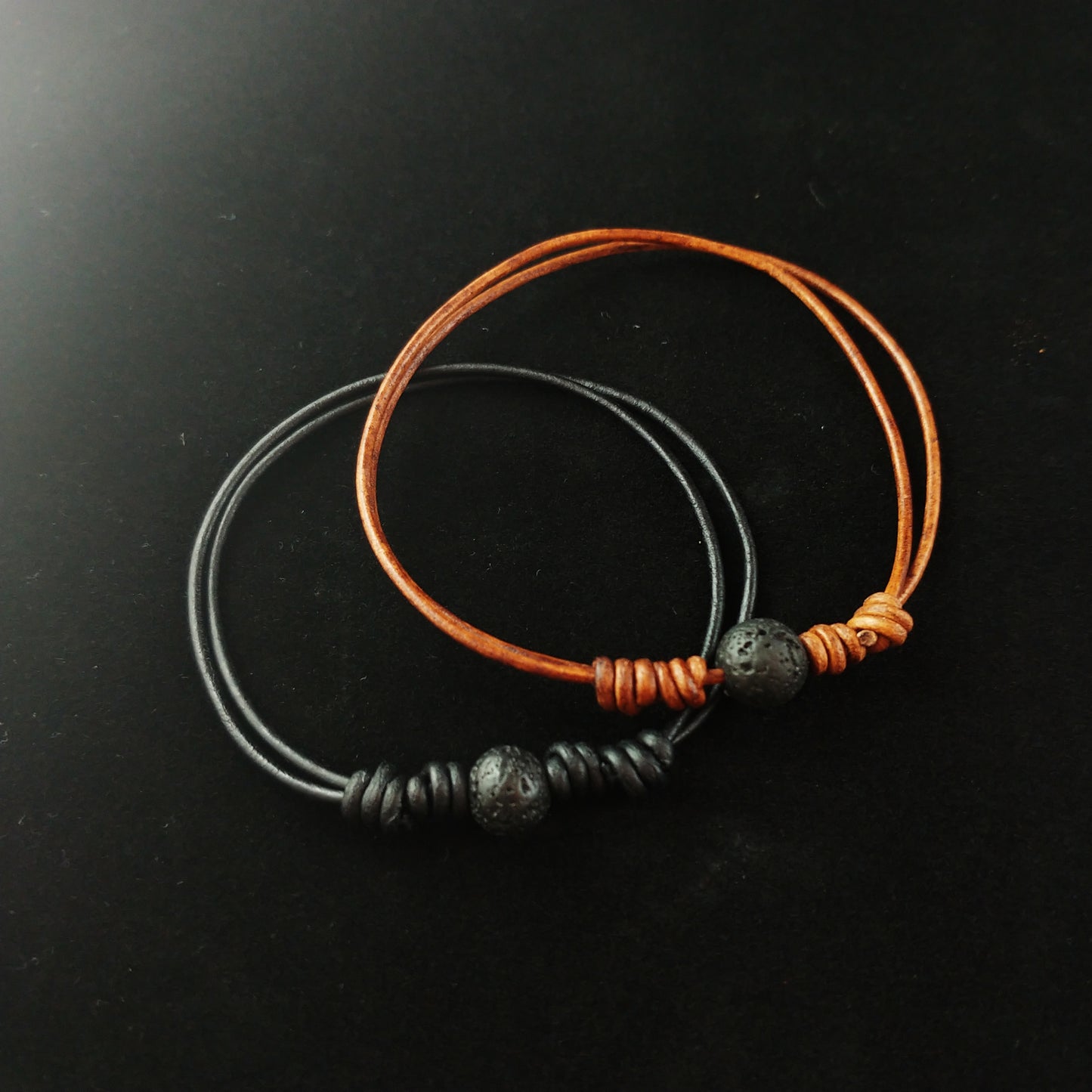 Thin leather bracelets sitting on a black display cloth. One bracelet has black leather and a center lava stone piece. The other bracelet has brown leather and a lava stone in the center.