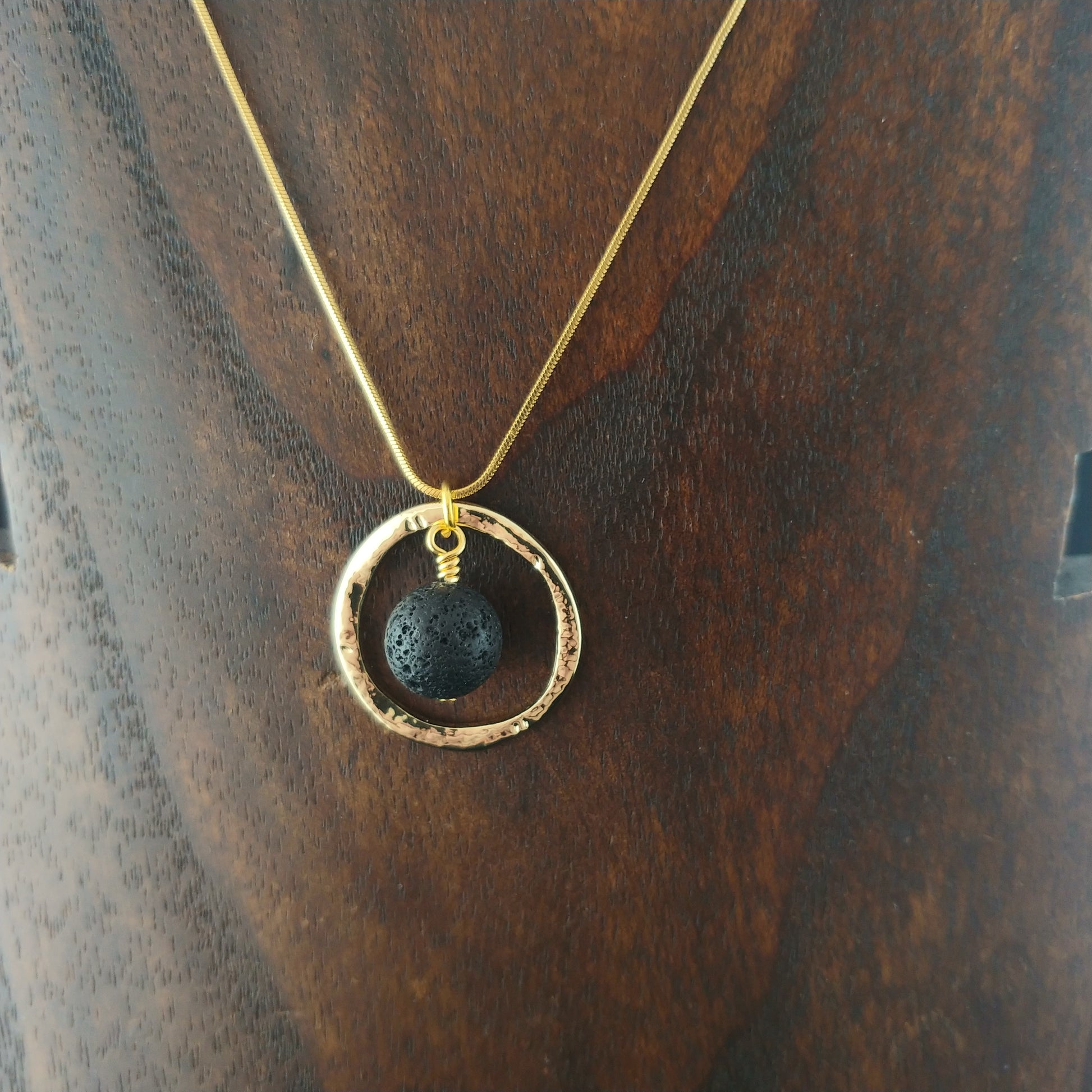 Golden circle necklace with a golden colored snake chain and a bright golden circle ring and in the middle of the rings there is a black lava bead. Necklace is being displayed on a wooden necklace display.