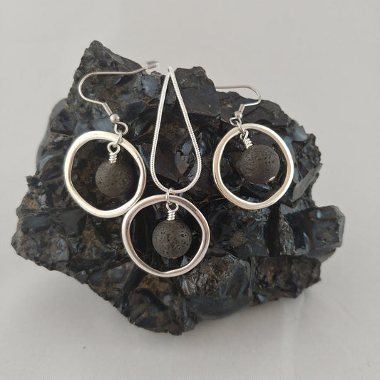 Jewelry set displayed on a dark black Obsidian stone display. There are two silver circle earrings with dangling lava beads in the center and between the earrings there is a necklace with a silver ring with dangling lava stone in the middle.