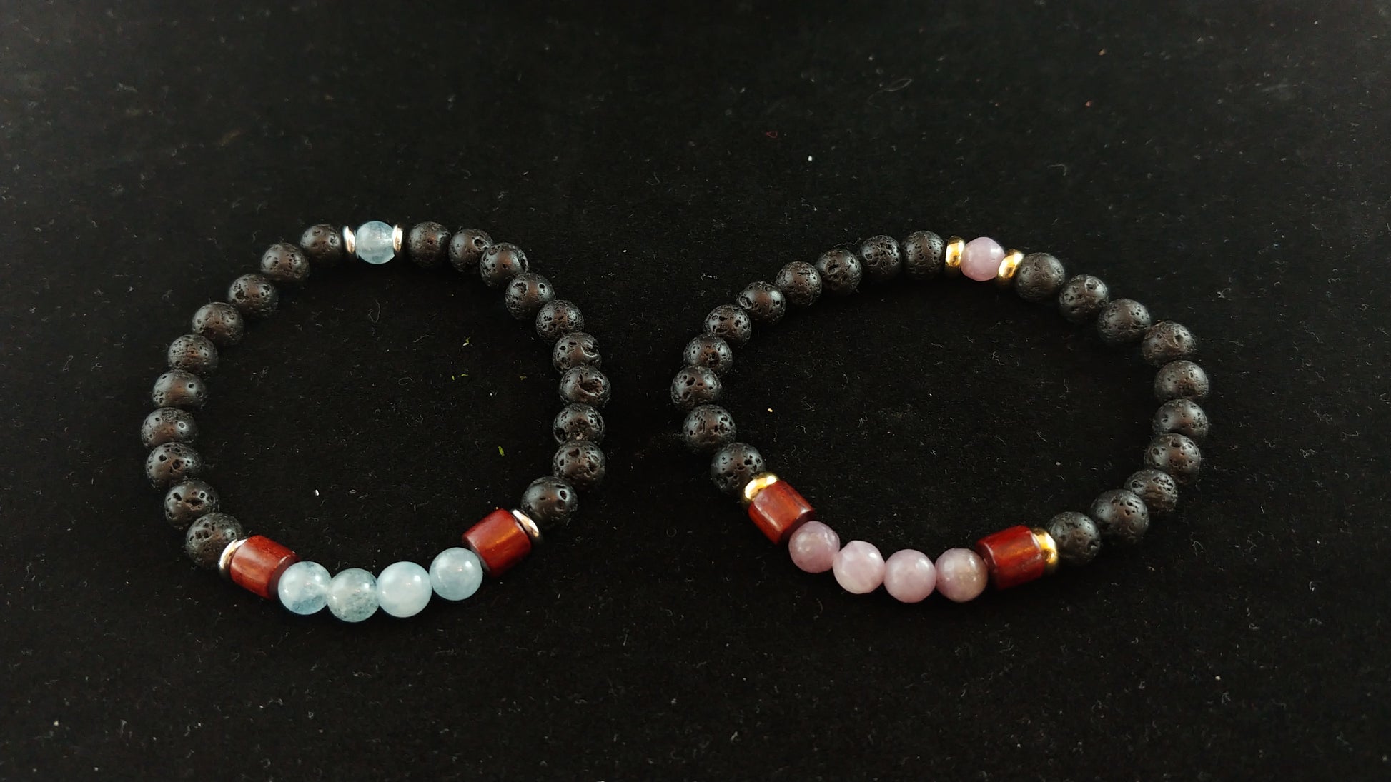 Two bracelets on a dark black background. One bracelet has bright blue aquamarine stones mixed with lava stones and wood and thin gold spacers. The other bracelet sits to the right and has the same golden and wooden spacers with lava and bright pink Aquamarine stones.