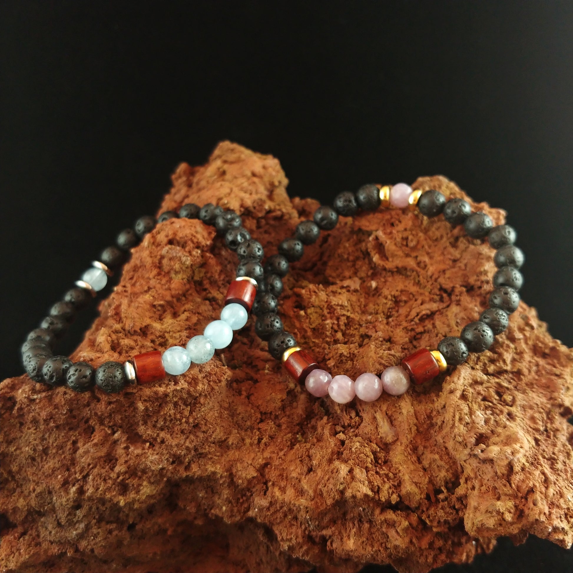 Two bracelets sitting on a red magma rock. One bracelet has bright blue aquamarine stones mixed with lava stones and wood and thin gold spacers. The other bracelet sits to the right and has the same golden and wooden spacers with lava and bright pink Aquamarine stones.
