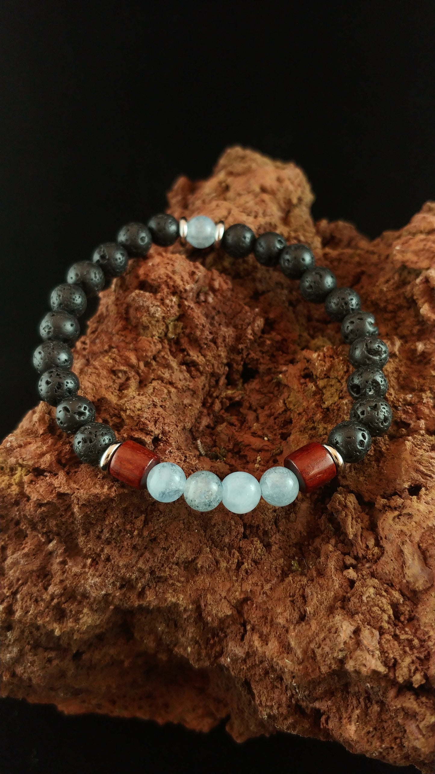 Bracelet being displayed on a dark red volcanic stone. Bracelet has mostly lava beads and bright blue Aquamarine beads. It also has thin golden colored spacers and larger wooden spacers between the lava and the aquamarine.