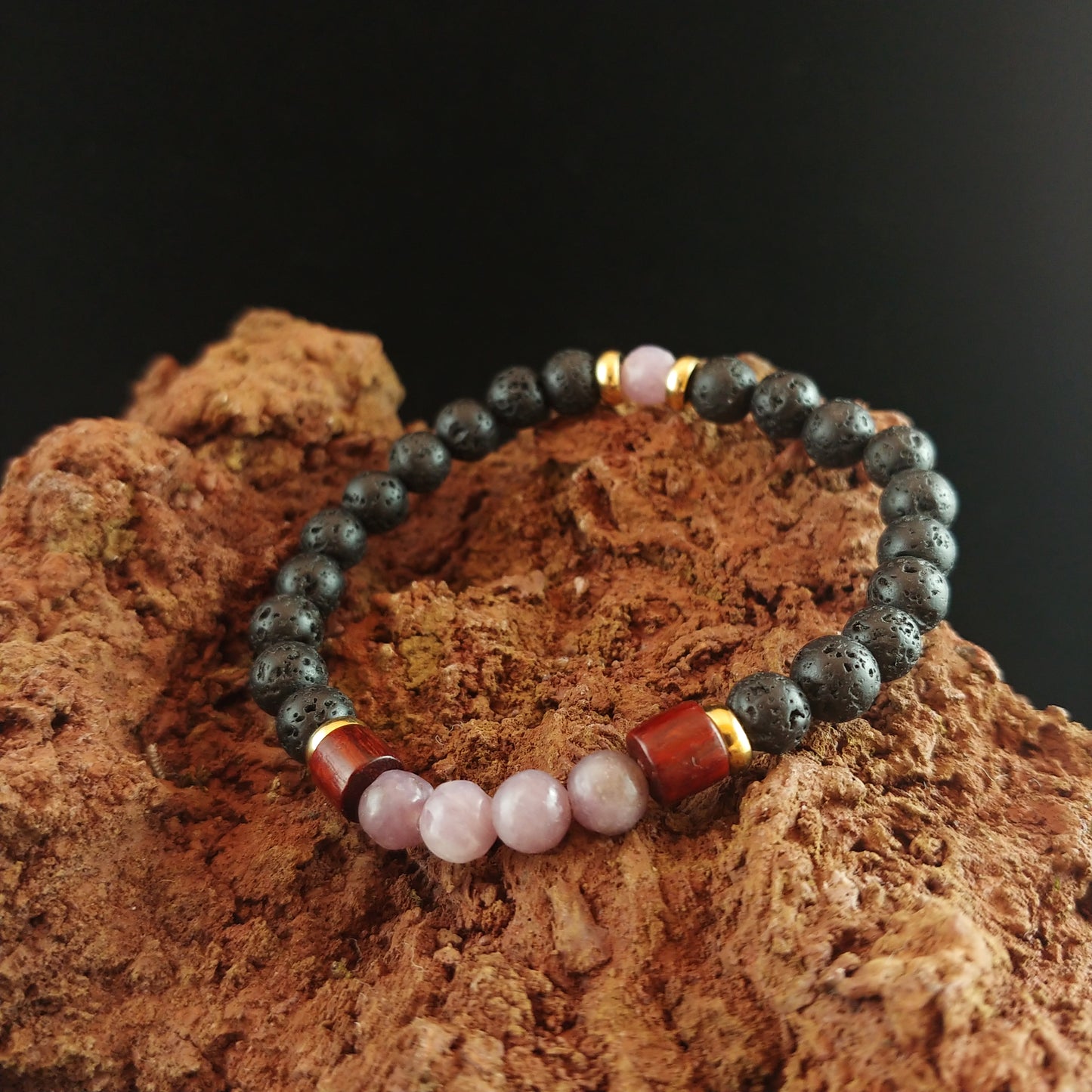 Bracelet sitting on a red lava stone. Bracelet is made of lava beads and a few bright pink Rose quartz, it also has thin golden colored spacers and larger wooden spacers between the lava and rose quartz.