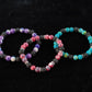 Three kids bracelets sitting on a dark black display cloth. The bracelet in the middle has a mix of bright red stones and lava beads. The bracelet on the left has bright purple stones and lava beads. The bracelet on the right has bright blue stones and lava beads.