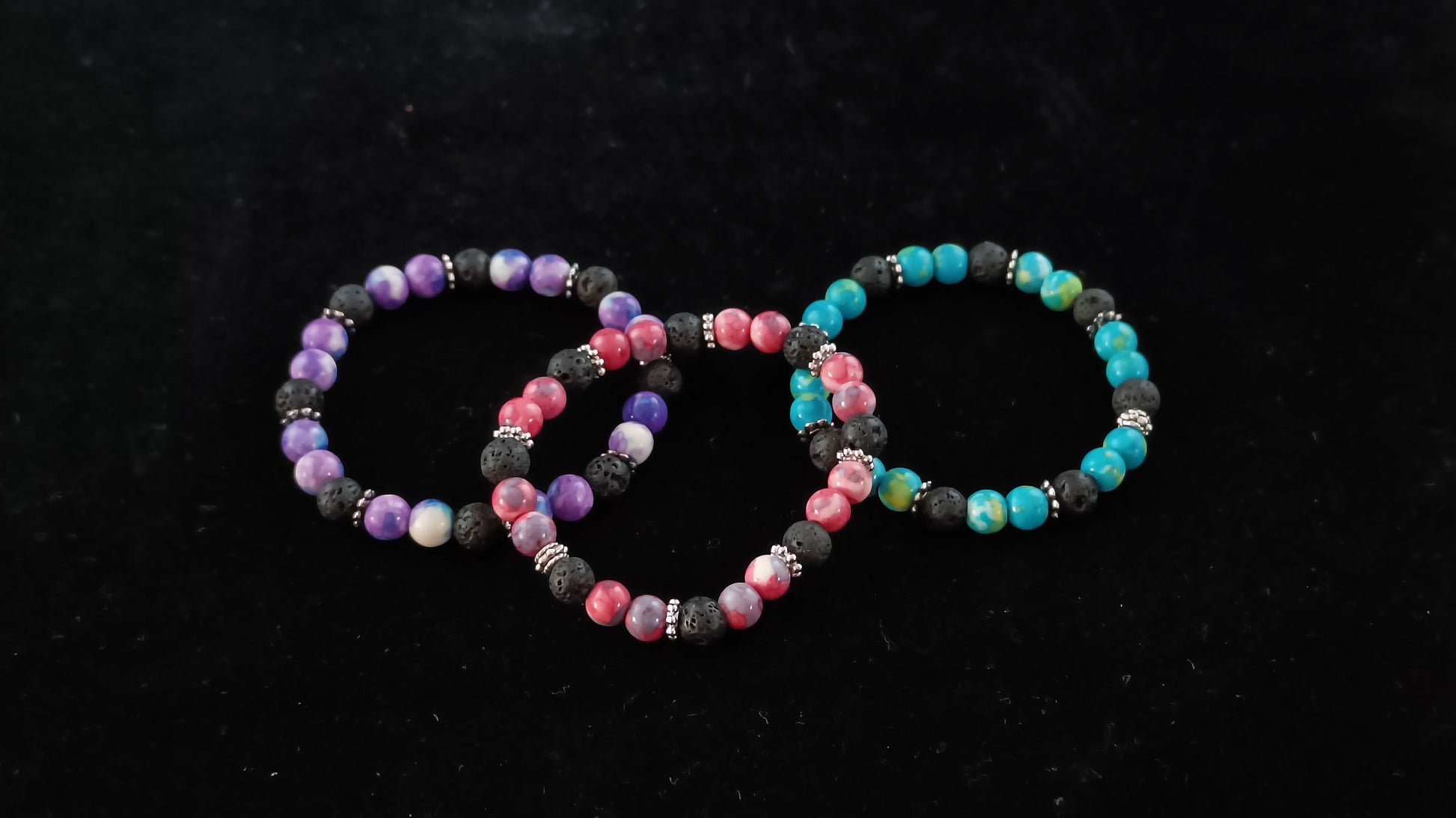 Three kids bracelets sitting on a dark black display cloth. The bracelet in the middle has a mix of bright red stones and lava beads. The bracelet on the left has bright purple stones and lava beads. The bracelet on the right has bright blue stones and lava beads.