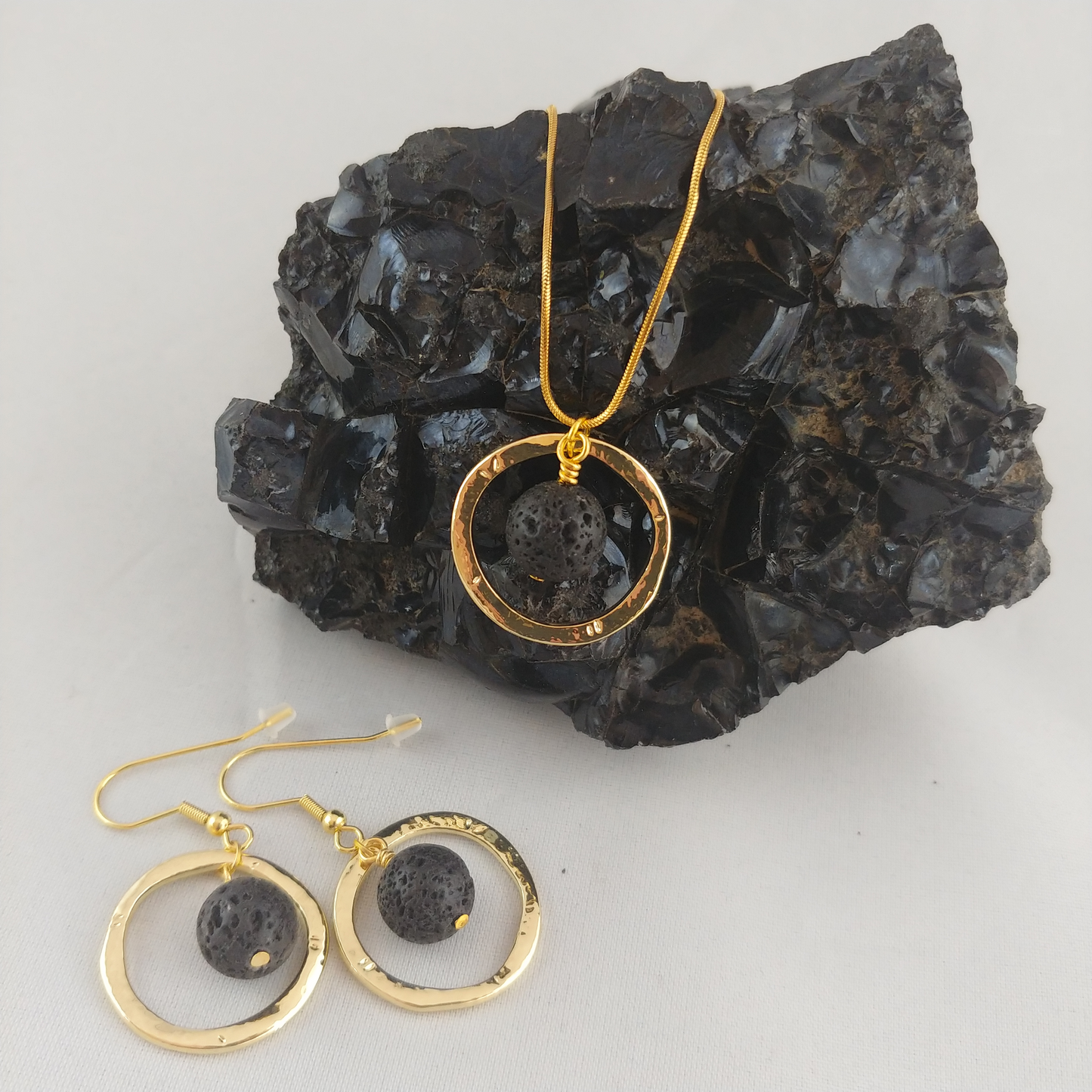 Set of golden colored Jewelry. A golden colored necklace with a golden colored ring with a lava stone in the middle sitting on a black Obsidian display. On the bottom left earrings with golden rings and in the middle lava beads.
