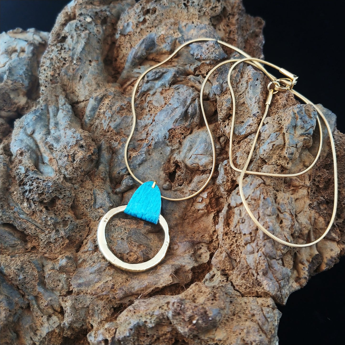 Necklace sitting on a black lava display. Necklace chain and circle center piece are golden color and the chain and circle are connected with icelandic fish leather that's brigh blue.