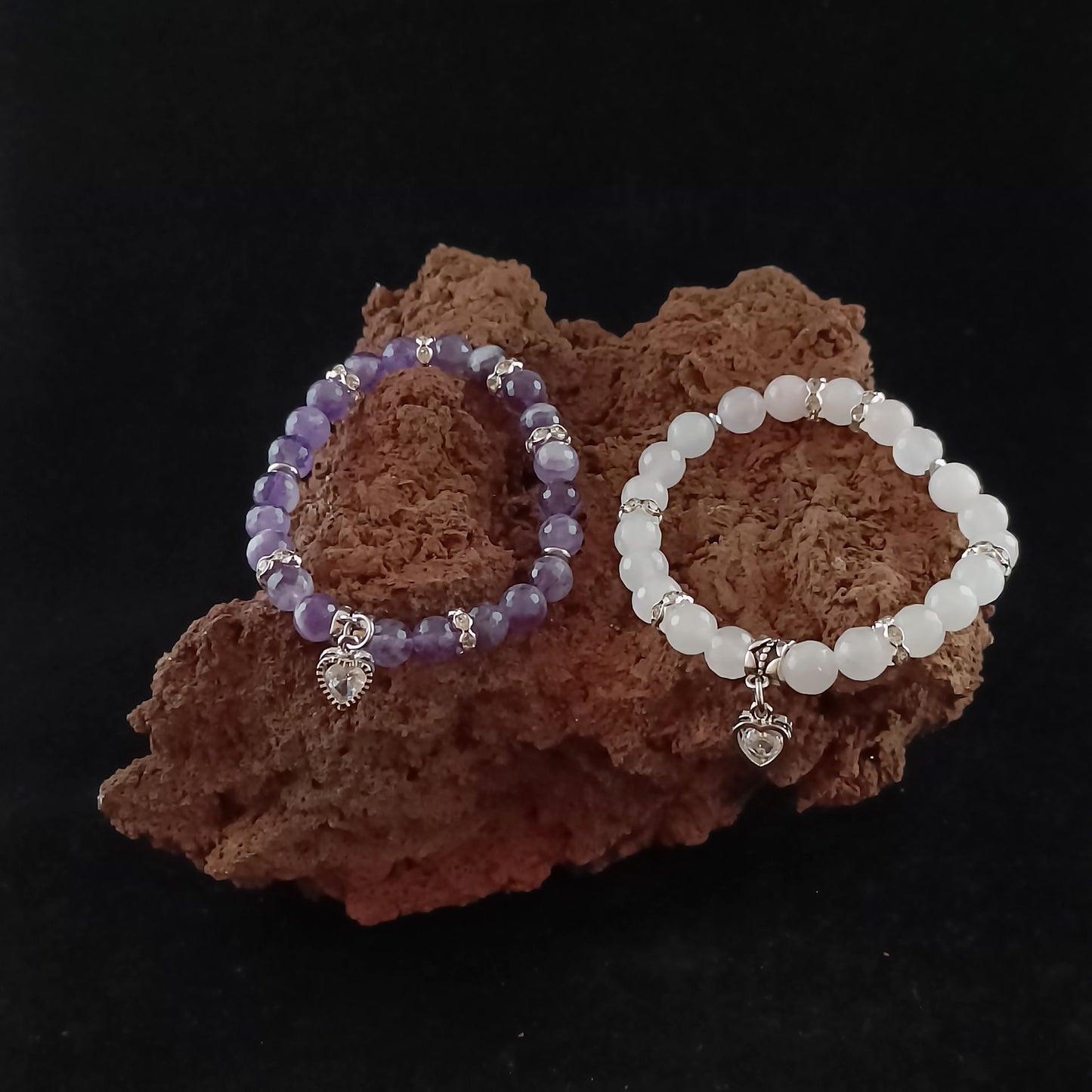 Two bright colored bracelets sitting on a dark red magma rock. On the left is a purple colored bracelet with a bright silver colored glass heart in the center. On the right is a bright pinkish beads bracelet with a heart centerpiece charm.