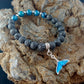 Lava Rock bracelet sitting on top af a magma rock display. The bracelet has dark black lava beads and brighter blue beads. In the center there is a bright blue whale tail charm.