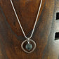 Silver colored circle necklace with a silver circle piece and in the middle is a black lava bead. Necklace is displayed on a dark brown wood necklace display.