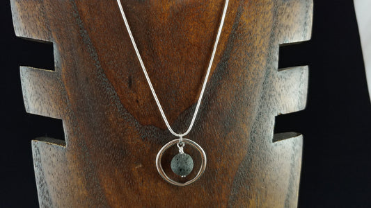 Silver colored circle necklace with a silver circle piece and in the middle is a black lava bead. Necklace is displayed on a dark brown wood necklace display.