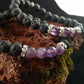 Two bracelets sitting on top of one another. The bottom bracelet has lava beads and 4 bright amethyst stones. The top bracelet has lava rock beads and three amethyst beads. The bracelet sit on a dark red lava rock.