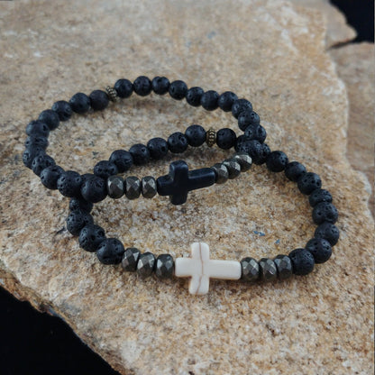 Two bracelets sitting on a display rock. One bracelet is sitting on top of the other. The bottom bracelet has small lava stones and a cross centerpiece. The other bracelet also has small lava stones but a dark black cross.