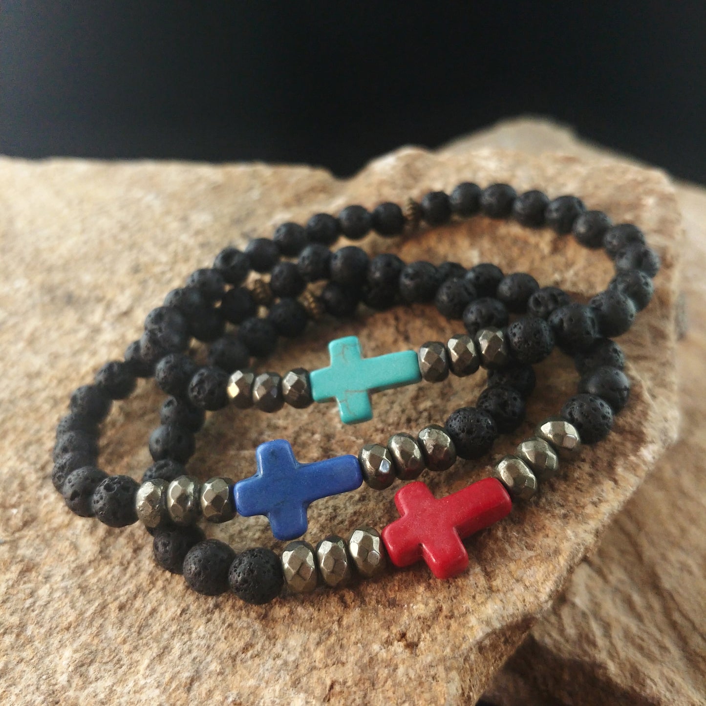 Three bracelets with cross center-piece sitting on top of one another. Below them is a bright brown stone display. All the bracelets have dark black lava beads, and bronze colored spacers beads. In the middle they all have a cross. The bottom bracelet has a red cross. The middle bracelet has a dark blue colored cross. The top bracelet has a turquoise colored cross.