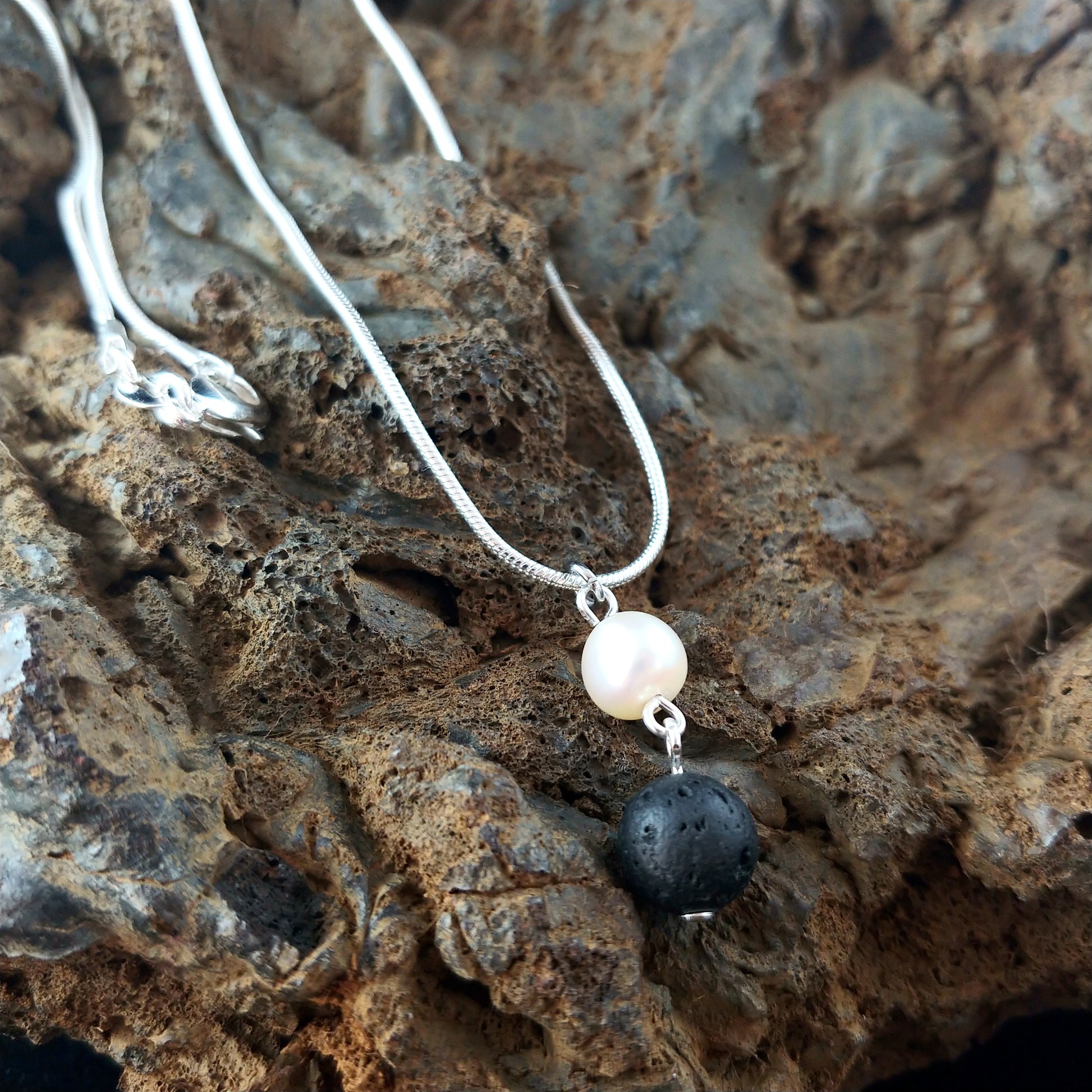 freshwater pearl necklace sitting on top of an old dark magma rock display. The necklace has a silver colored snake chain. The pendant is made from lava and freshwater pearl.