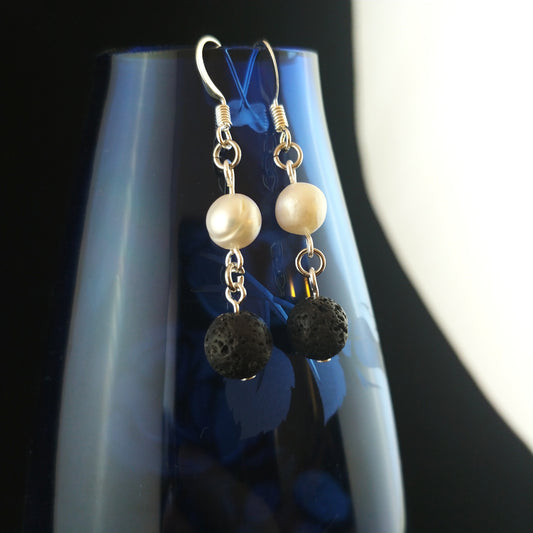 Earrings with black 8mm lava beads on the bottom and above them bright freshwater pearls. Earrings are displayed on a blue transparent flower display.