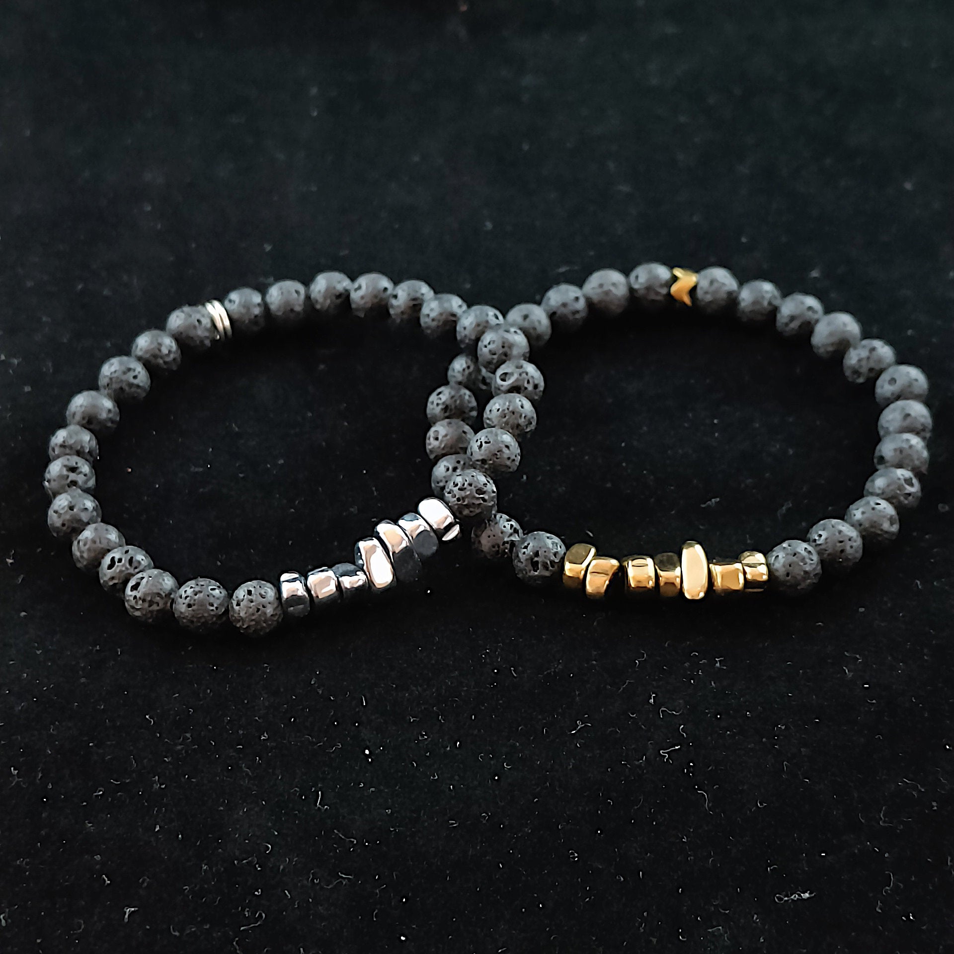 two bracelets sitting on a black cloth. Both bracelets are made with lava beads and in the center are colored shards pieces. On the right bracelet they're colored golden but on the left bracelet they're silver colored.
