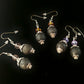 Three earrings sitting on a dark black display cloth. All the earrings have a black lava bead with a silver colored cap. On top of each earrings is a colored glass piece. On the far right the glass is purple, in the middle the glass is orange and one the left side earrings the glass is clear