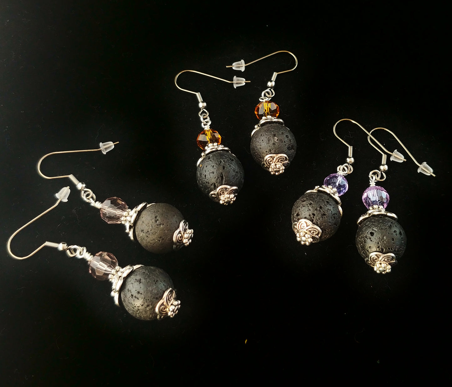 Three earrings sitting on a dark black display cloth. All the earrings have a black lava bead with a silver colored cap. On top of each earrings is a colored glass piece. On the far right the glass is purple, in the middle the glass is orange and one the left side earrings the glass is clear
