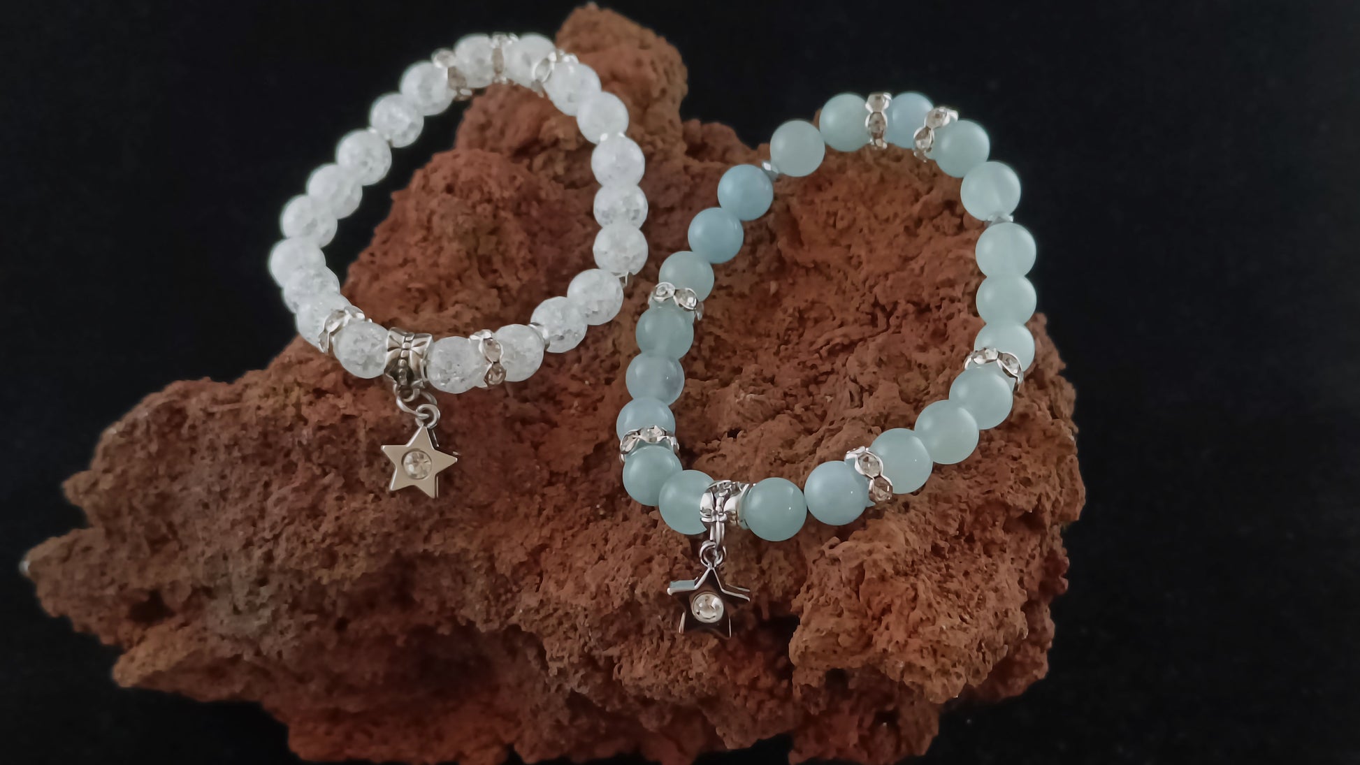 Two bright bracelets sit togather on a red magma display stone. On the left is a bracelet made from Cracked crystal beads and has a star center piece charm. On the right is a bracelet with bright blue aquamarine beads and a star center-piece